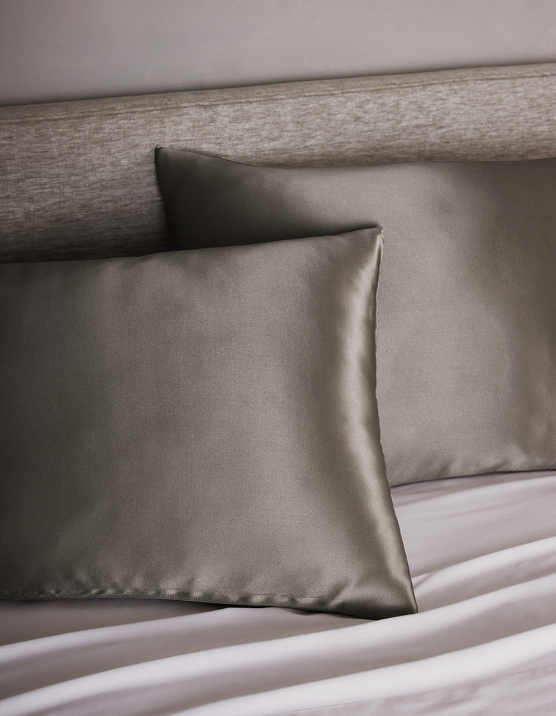 Two Cozy Earth Silk Pillowcases are placed on a bed with a light gray upholstered headboard and white sheets, adding a modern and elegant touch.