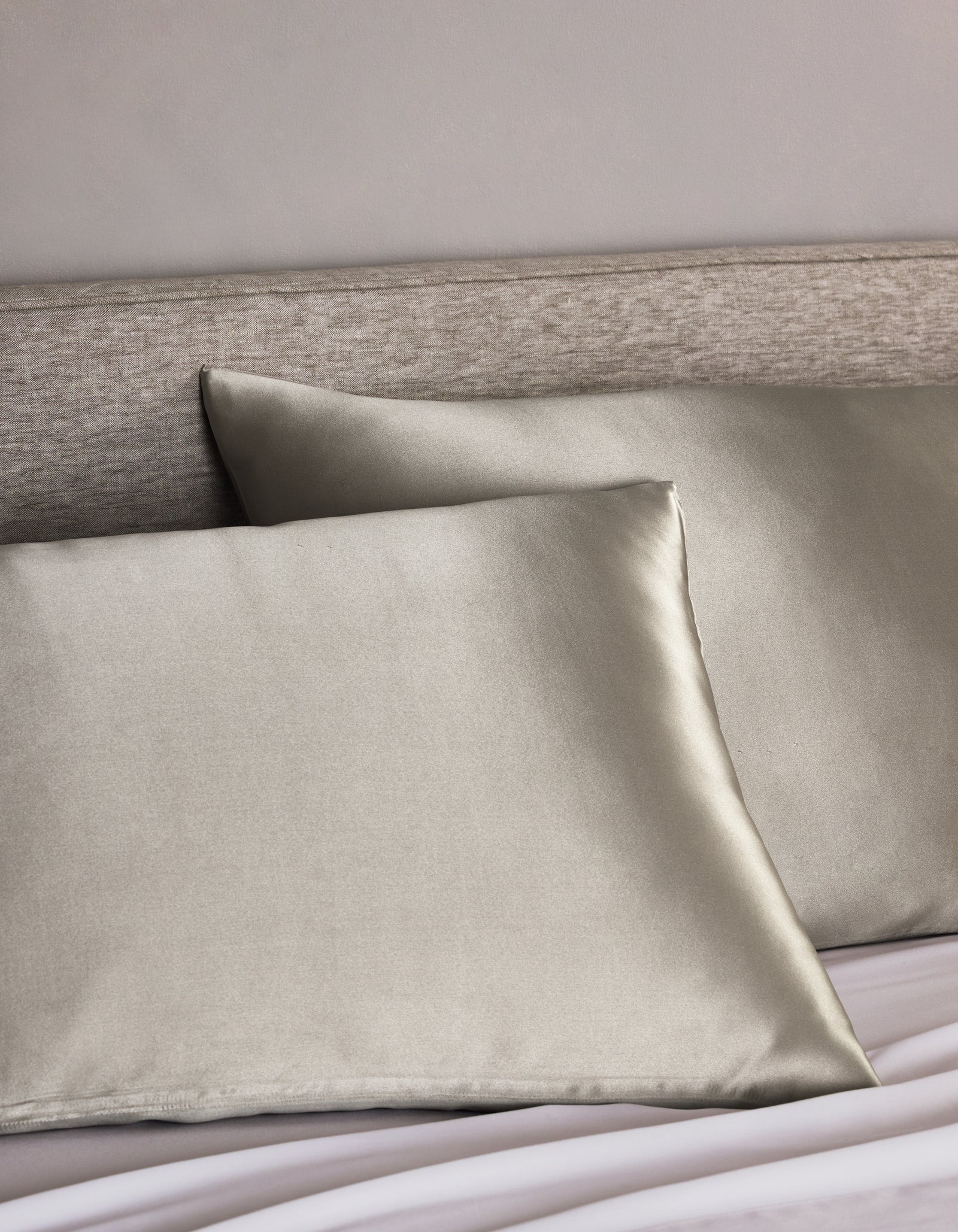 Two Cozy Earth Silk Pillowcases in light gray are arranged on a bed with a light beige upholstered headboard, adding a smooth, shiny, and luxurious touch to the bedding.