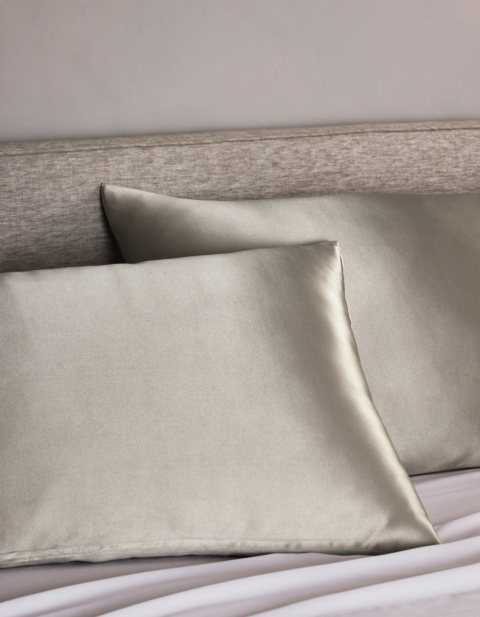 Two Cozy Earth Silk Pillowcases in light gray are arranged on a bed with a light beige upholstered headboard, adding a smooth, shiny, and luxurious touch to the bedding.