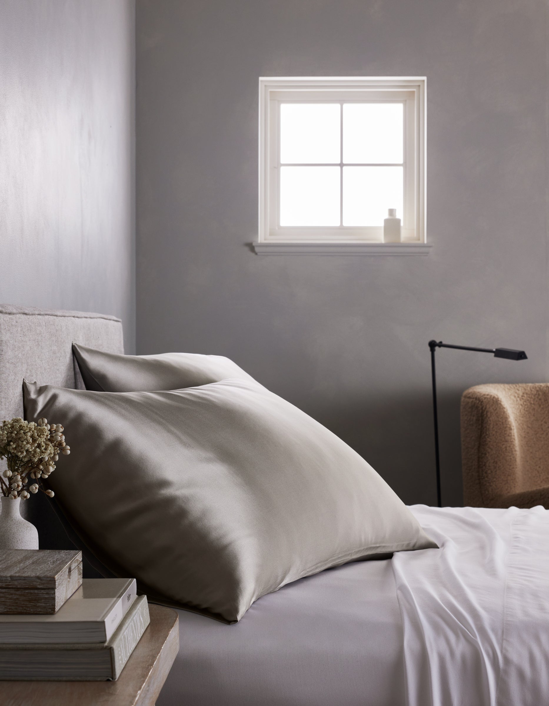 A neatly made bed features large, smooth Cozy Earth Silk Pillowcases in silver-gray and crisp white sheets beside a small table with books and flowers. A soft chair, lamp, and window on a gray wall complete the minimalist bedroom.|Color:Dove Grey