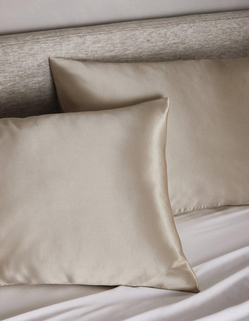 Two Cozy Earth Silk Pillowcases in light beige are displayed on a bed with a matching headboard and white sheets, creating a soft and elegant bedding arrangement.