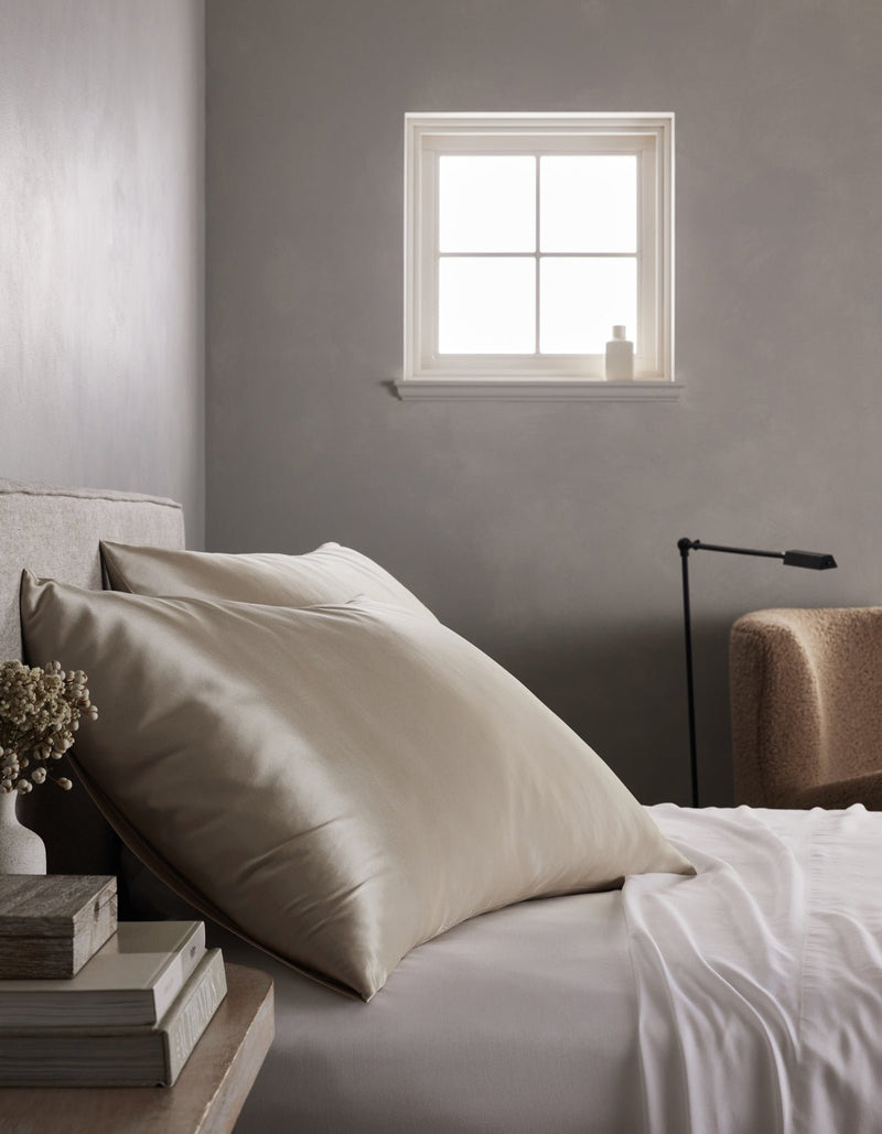 A neatly made bed features large cream Cozy Earth Silk Pillowcases and a white sheet beside a small window. A stack of books and dried flowers sit on the bedside table, with a beige armchair and black lamp in the background.