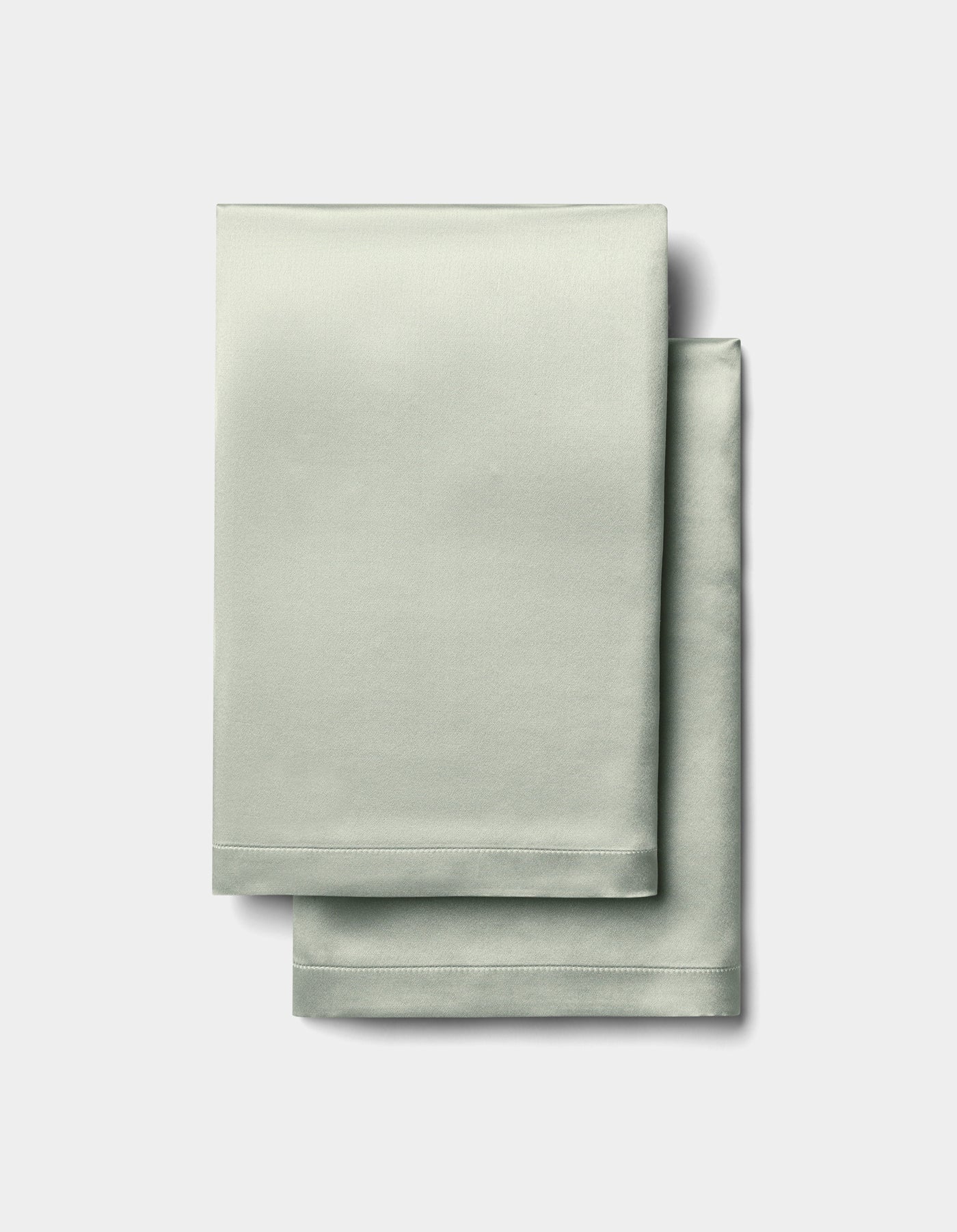 Two neatly folded Cozy Earth Silk Pillowcases in light sage green, featuring a smooth, satin-like texture, are stacked on a plain white background.|Color:Laurel