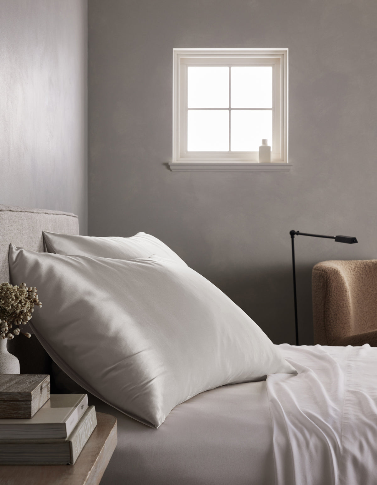 A neatly made bed with Cozy Earth Silk Pillowcases sits beside a stack of books and dried flowers. Soft light streams through a small window, while a beige armchair and black reading lamp are visible in the background.