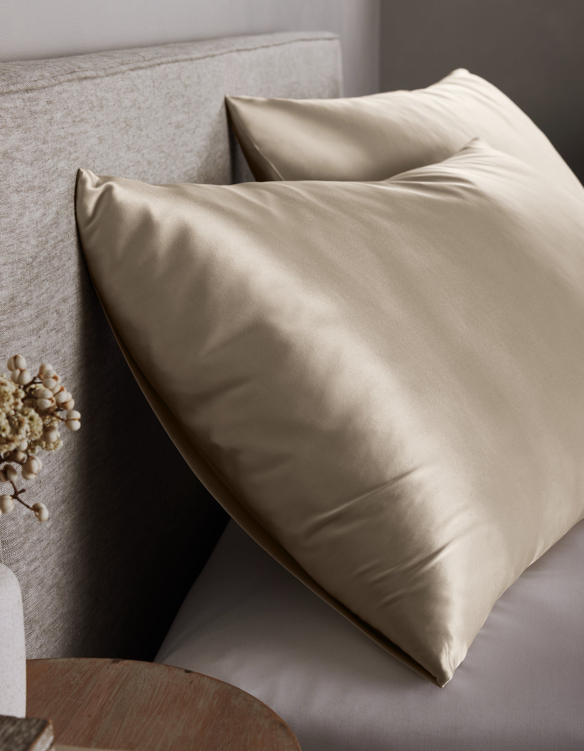 Two large, shiny Cozy Earth Silk Pillowcases in champagne rest against a light upholstered headboard on the bed, with a small round wooden nightstand and a vase of dried flowers nearby.