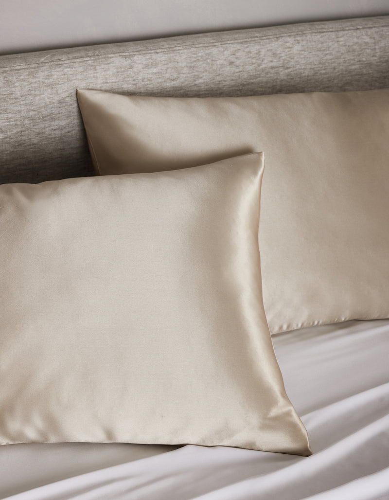 Two Cozy Earth Silk Pillowcases in beige are displayed on a bed with a light gray upholstered headboard and white sheets.