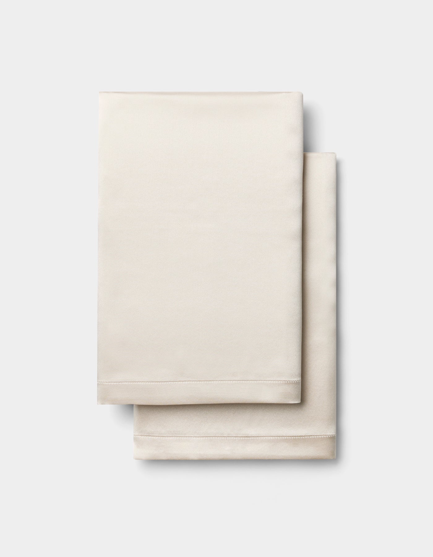 Two neatly folded, light beige Silk Pillowcases by Cozy Earth are stacked on a plain, light gray background.