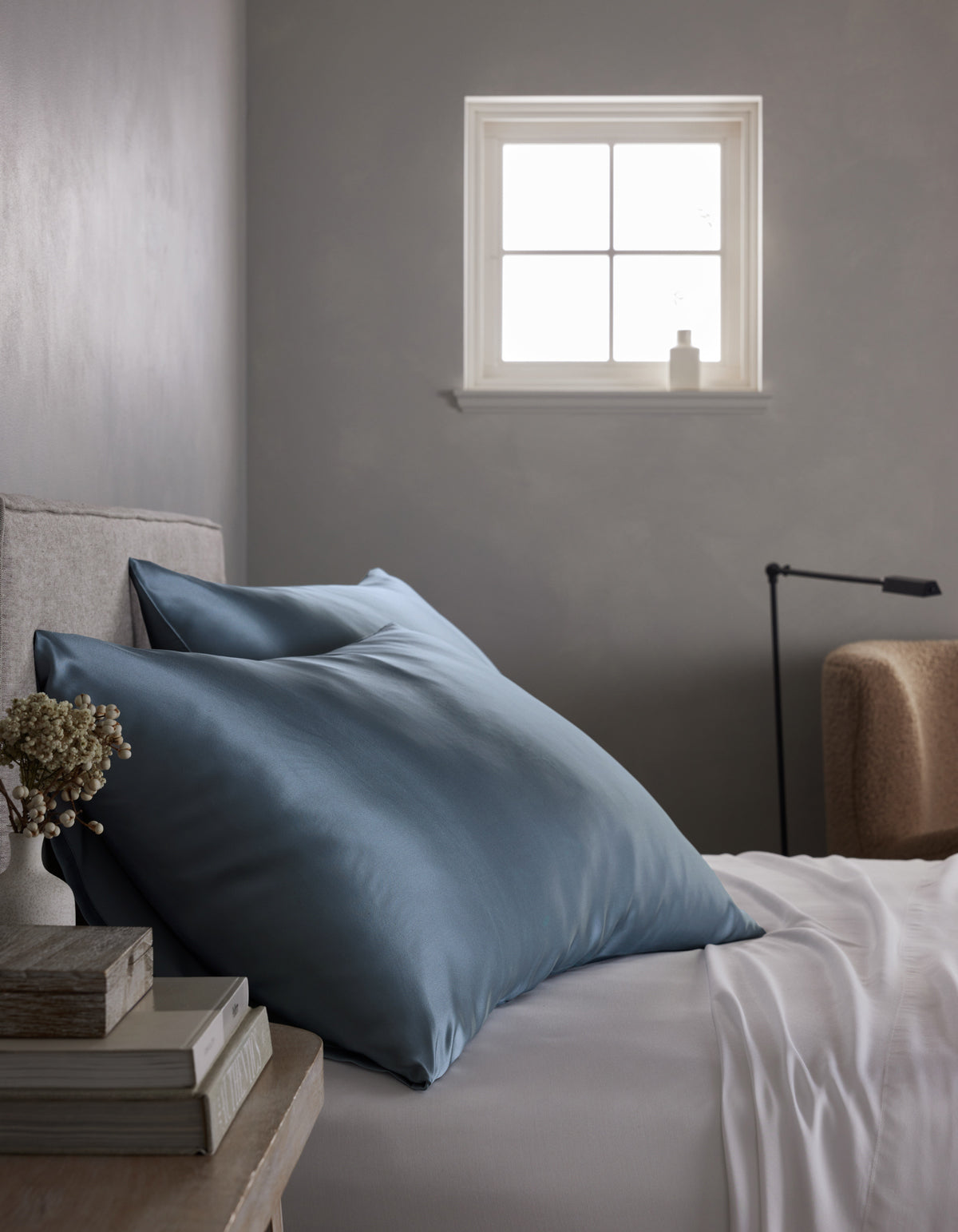 A neatly made bed with Cozy Earth Silk Pillowcases and a white sheet sits beside a small window in a softly lit, minimal bedroom. A stack of books and a small vase are arranged on the bedside table.