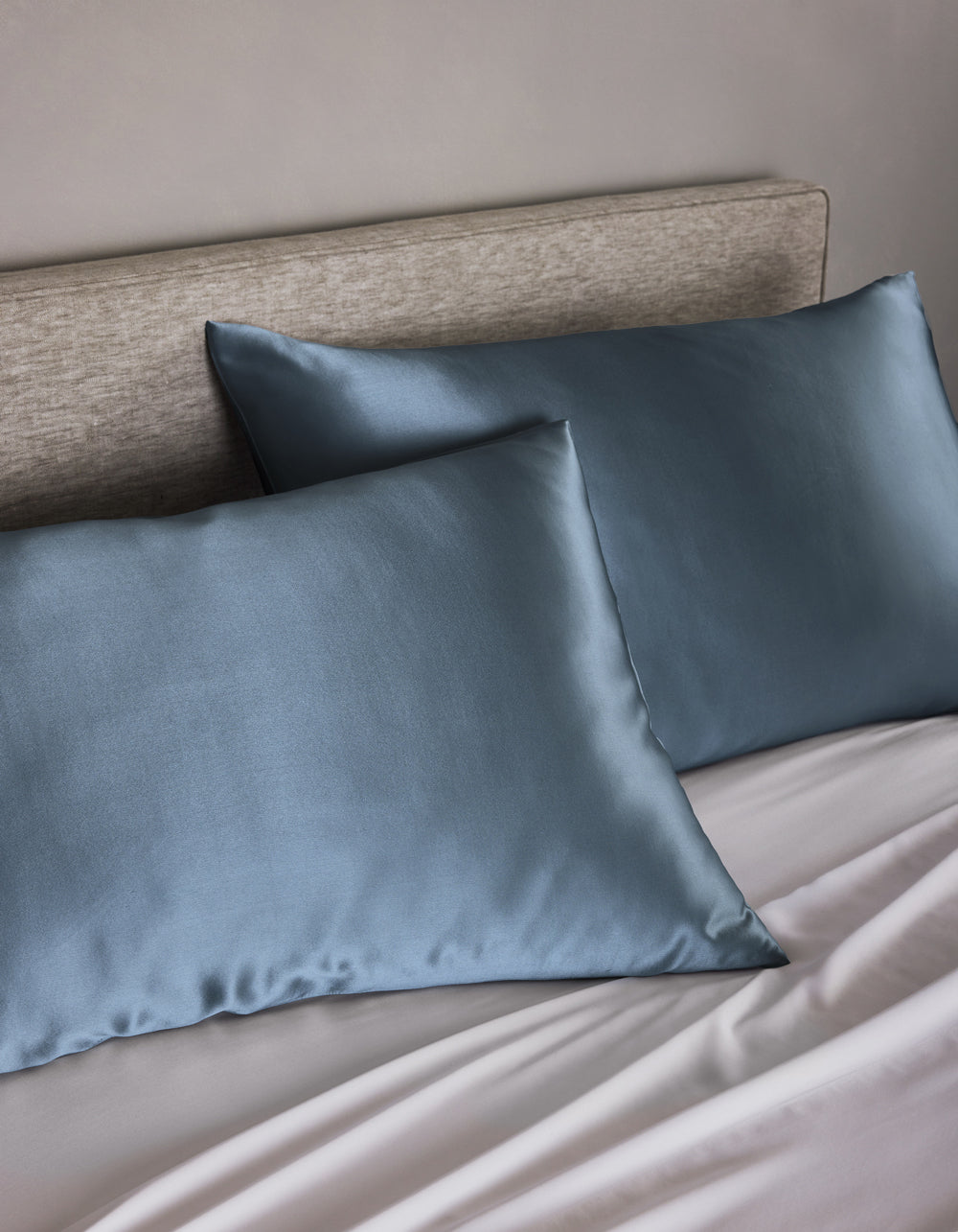 Two Cozy Earth Silk Pillowcases in blue rest on pillows against a light beige upholstered headboard, complemented by light gray sheets. The look is clean and minimalistic.