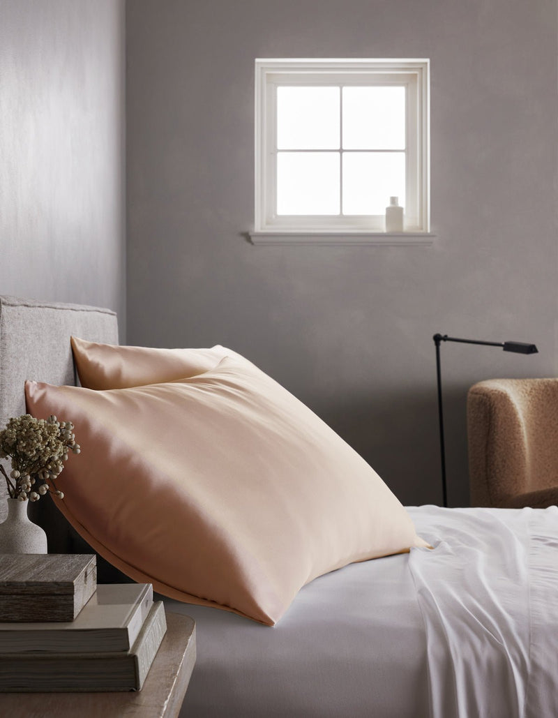 A neatly made bed features large, light-colored Silk Pillowcases by Cozy Earth beside a small table with stacked books and dried flowers, under a sunlit window. In the background are a beige armchair and a floor lamp.