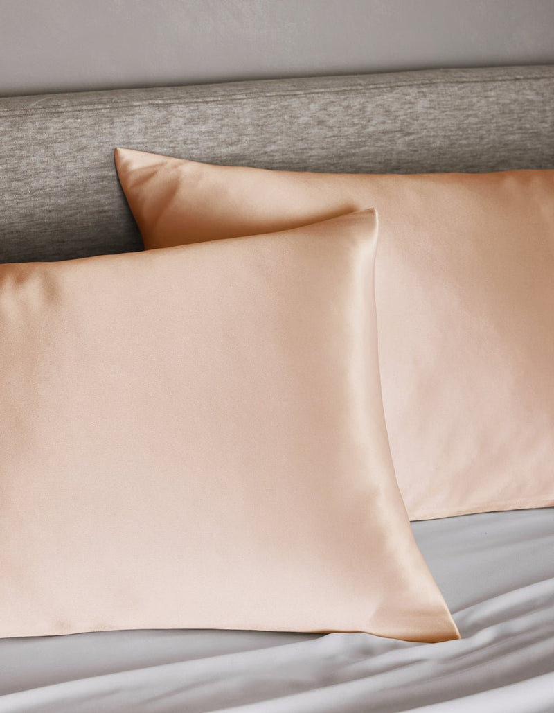 Two Cozy Earth Silk Pillowcases in peach rest on a bed with a light gray upholstered headboard and white sheets.