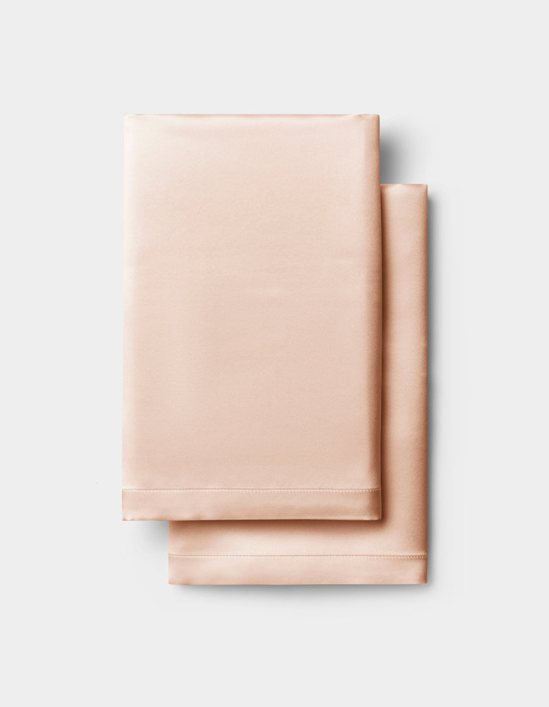 Two neatly folded Cozy Earth Silk Pillowcases in light peach are stacked on a light gray background.