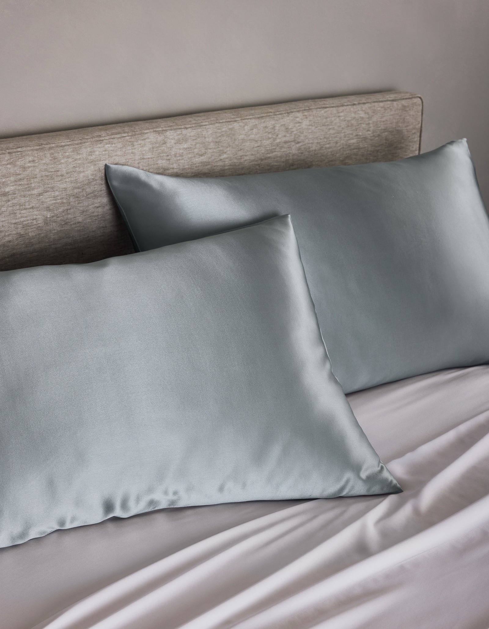 Two Cozy Earth Silk Pillowcases in silver-gray rest on a beige-upholstered headboard bed with matching light gray sheets, creating a soft and elegant look.