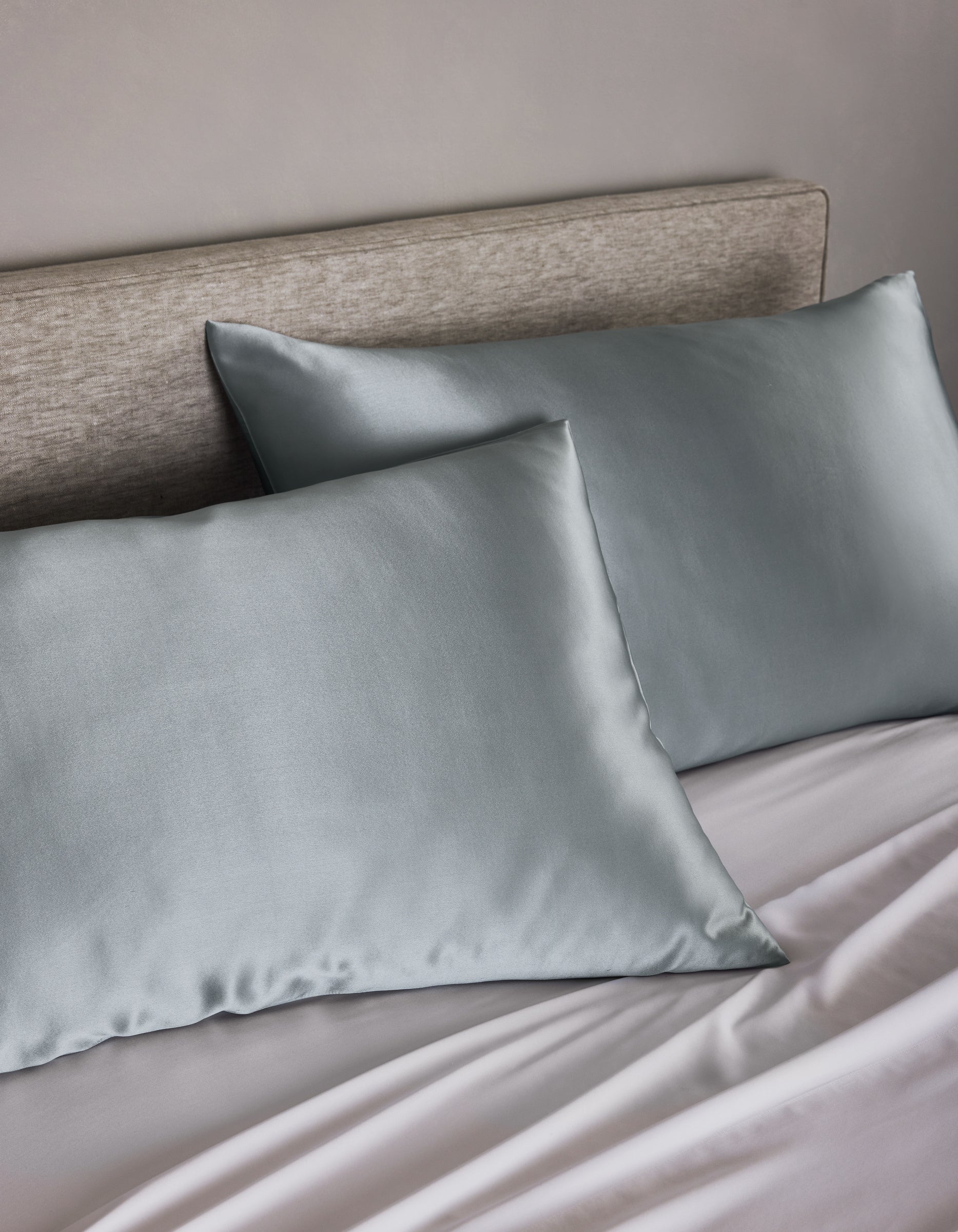 Two Cozy Earth Silk Pillowcases in silver-gray rest on a beige-upholstered headboard bed with matching light gray sheets, creating a soft and elegant look.