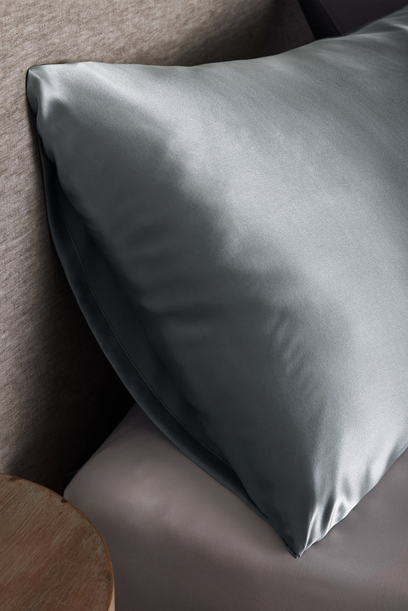 A Cozy Earth Silk Pillowcase in smooth, shiny gray sits on a bed with light gray sheets beside a wooden nightstand and a textured fabric headboard.