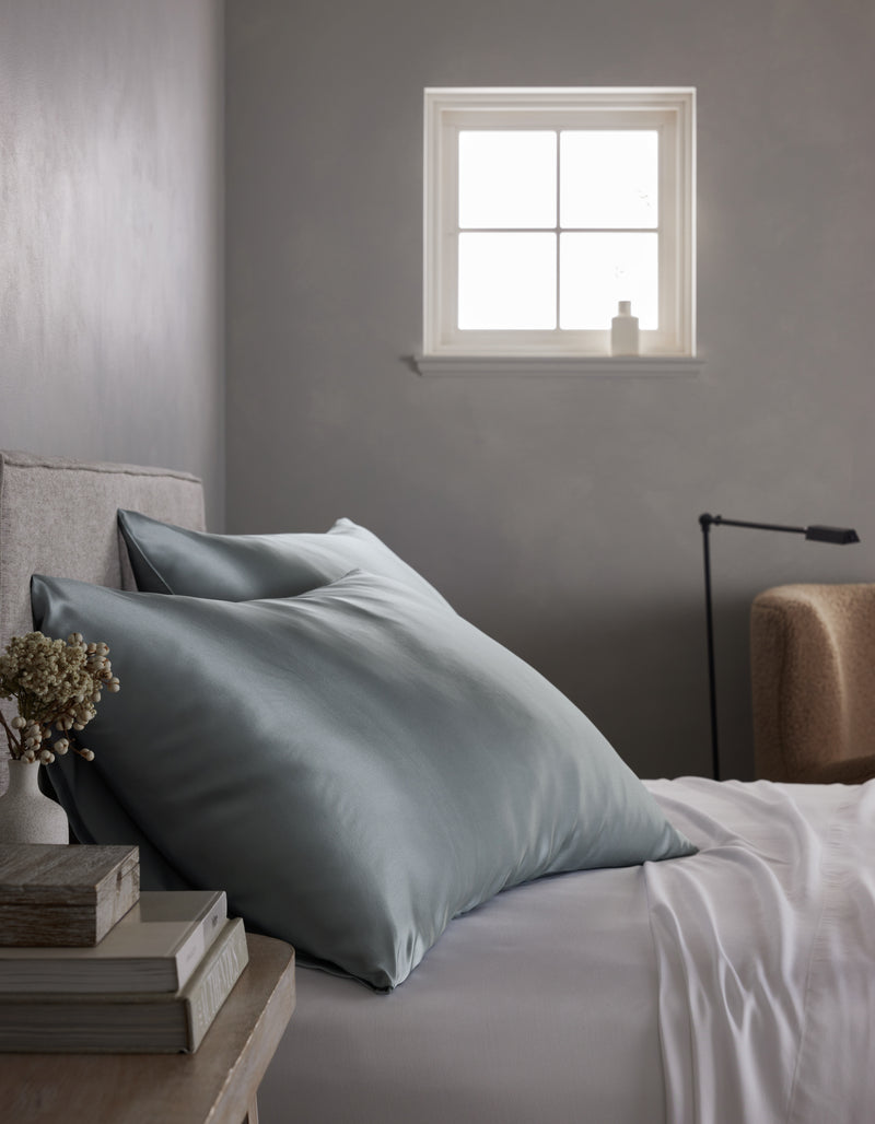 A neatly made bed with Cozy Earth Silk Pillowcases and white sheets sits by a small wooden nightstand holding books, a vase of dried flowers, and a window letting in soft natural light.