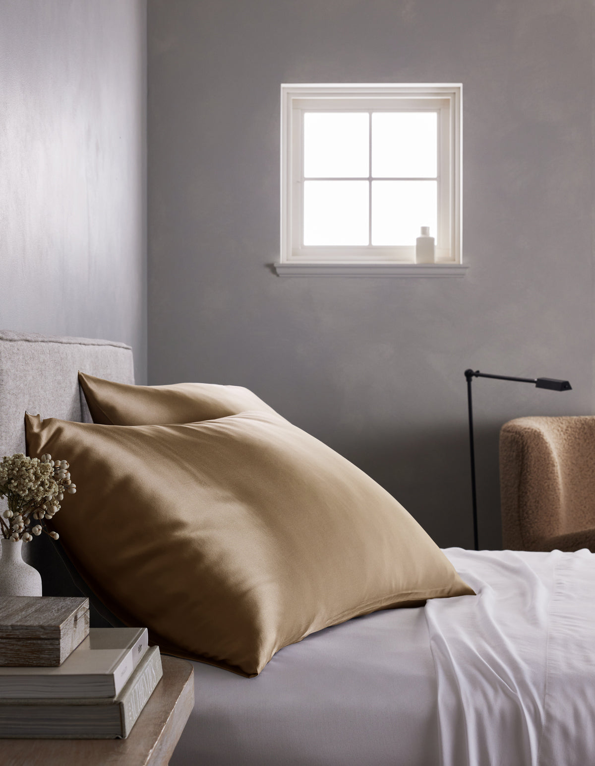A neatly made bed features a Cozy Earth Silk Pillowcase, crisp white sheets, and a stack of books on the bedside table, all set against a gray wall with soft natural light streaming in through the window.