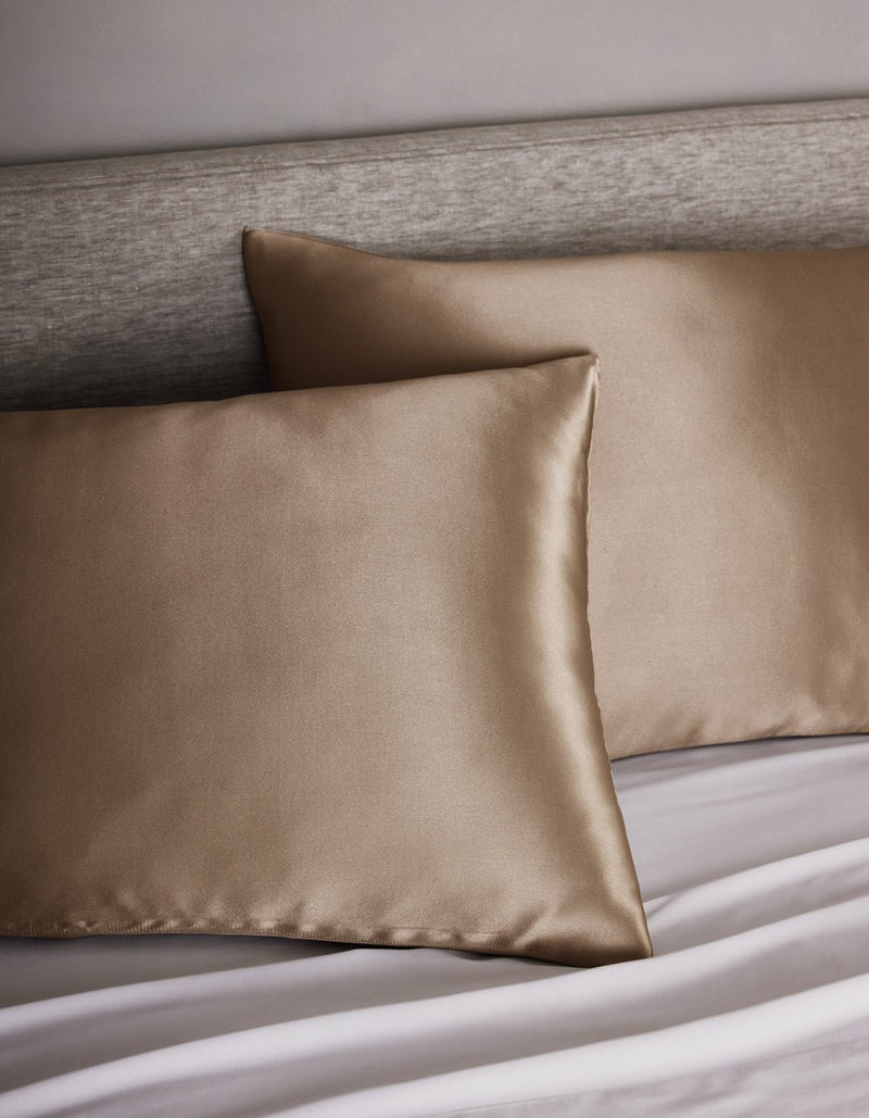 Two Cozy Earth Silk Pillowcases rest on a bed with a light gray upholstered headboard and white sheets.