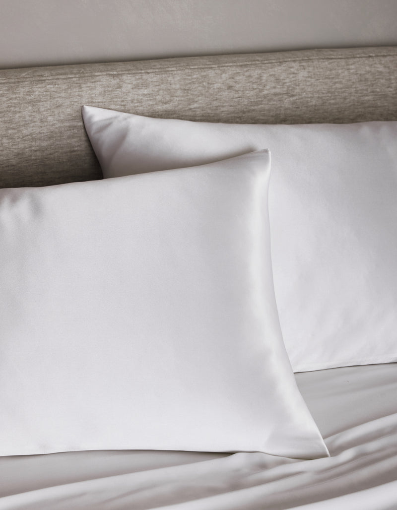 Two Cozy Earth Silk Pillowcases rest on a neatly made bed with a light gray upholstered headboard, creating a clean and minimalistic bedroom atmosphere.