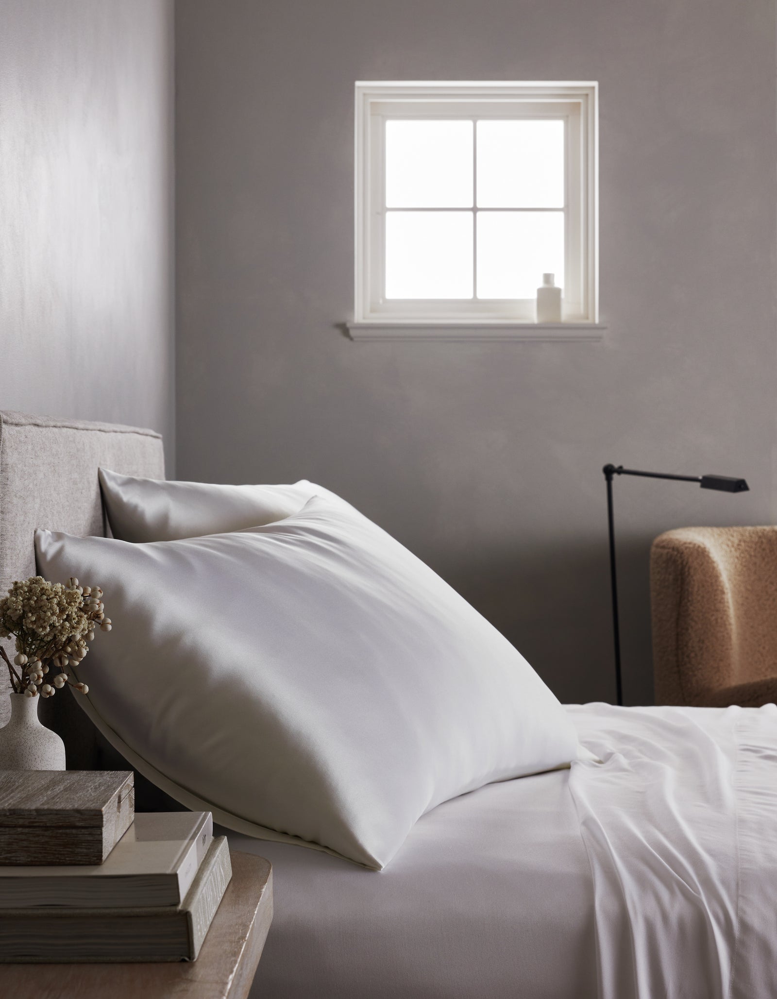 A neatly made bed features a Cozy Earth Silk Pillowcase, with white sheets beside a nightstand holding books and dried flowers. Natural light streams in from the window, illuminating a beige armchair and black lamp in the background.