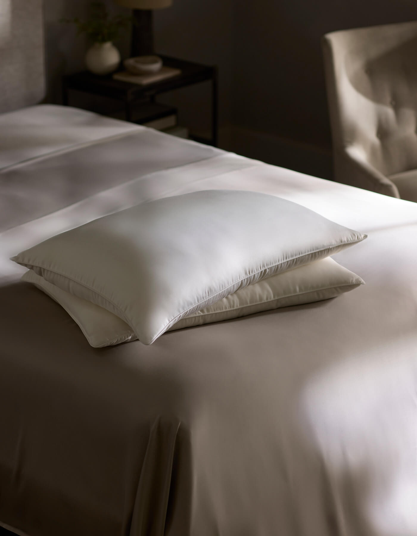 Two Silk Insert Bundle pillows by Cozy Earth are stacked on a bed with smooth taupe bedding. In the blurred background, there’s a bedside table with a plant and books, plus a beige upholstered chair.