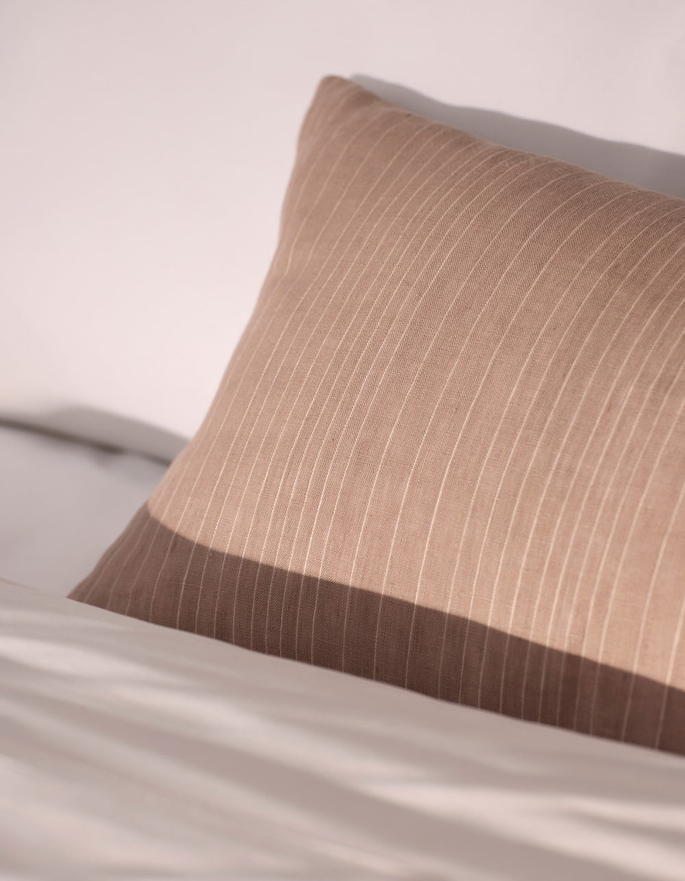 The Cozy Earth Striped Throw Pillow, in beige with subtle vertical stripes, rests against white bedding in soft natural light.