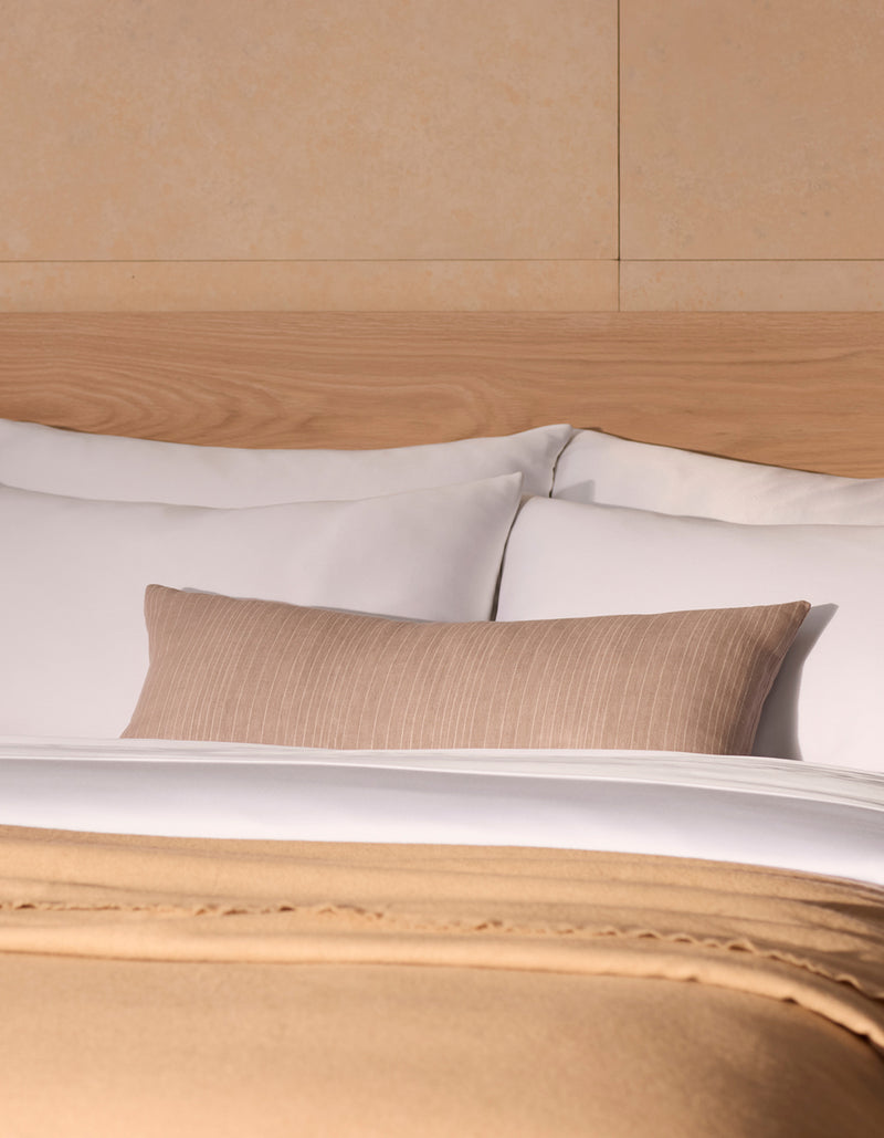 A neatly made bed with white pillows, a Cozy Earth Striped Throw Pillow, a light wooden headboard, and a beige blanket folded at the foot of the bed.
