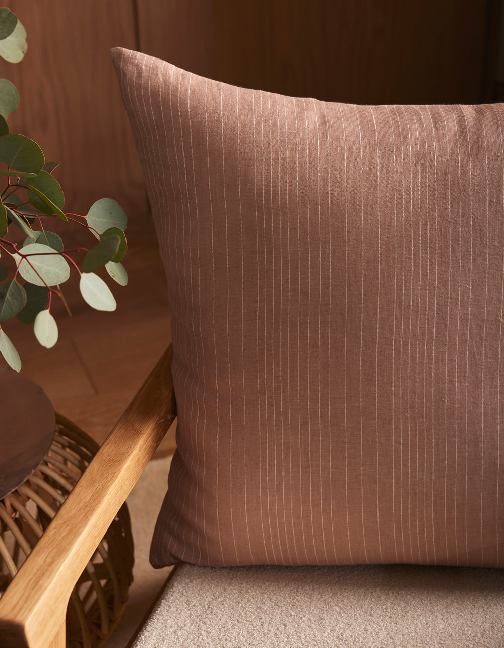 The Cozy Earth Striped Throw Pillow features subtle vertical white pinstripes on brown fabric, shown on a light cushioned chair beside a wooden table with eucalyptus branches.