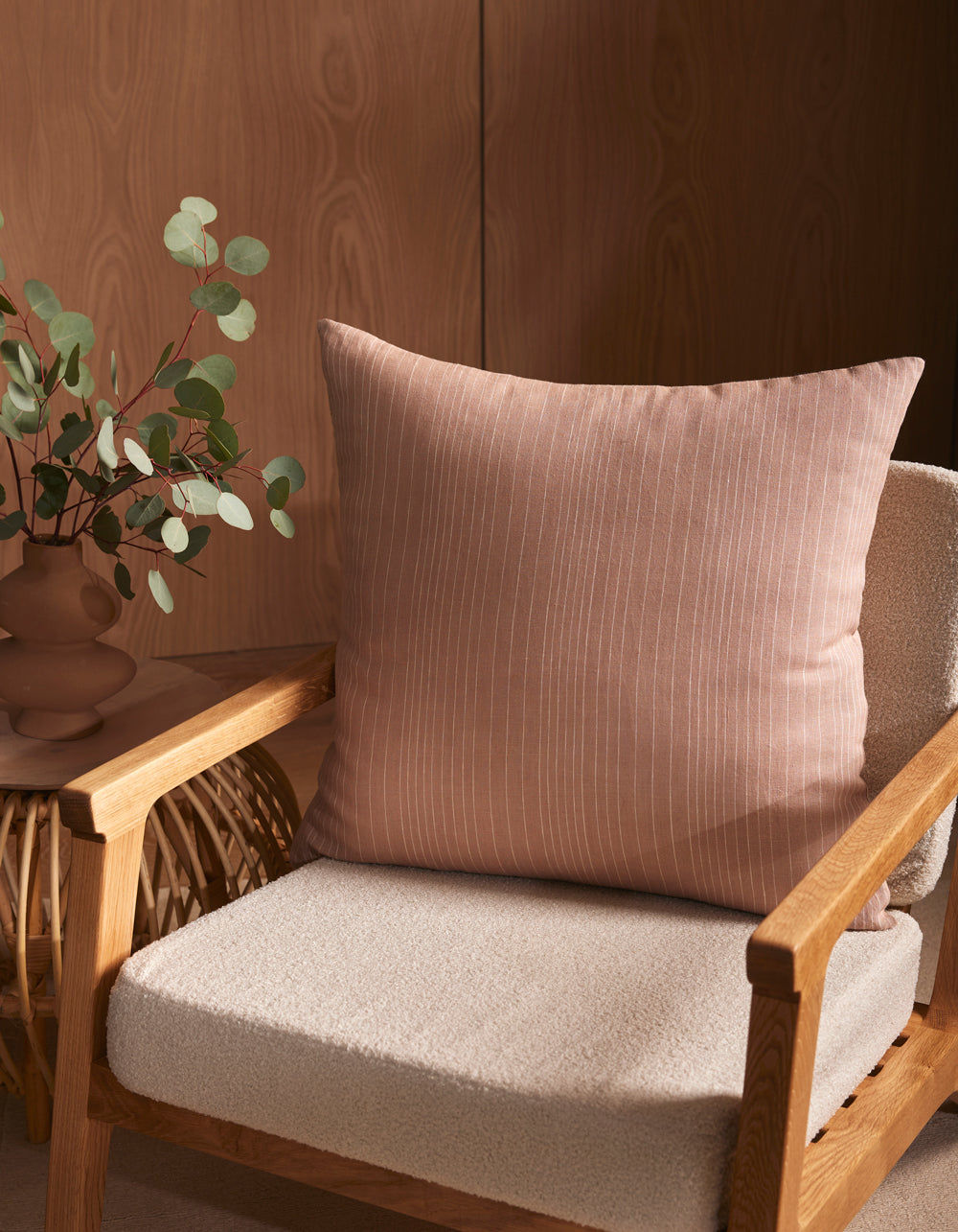A soft beige armchair with a textured seat cushion features the Striped Throw Pillow by Cozy Earth in pale pink. Beside the chair, a wicker side table holds a vase with eucalyptus branches, all set against a wooden wall.