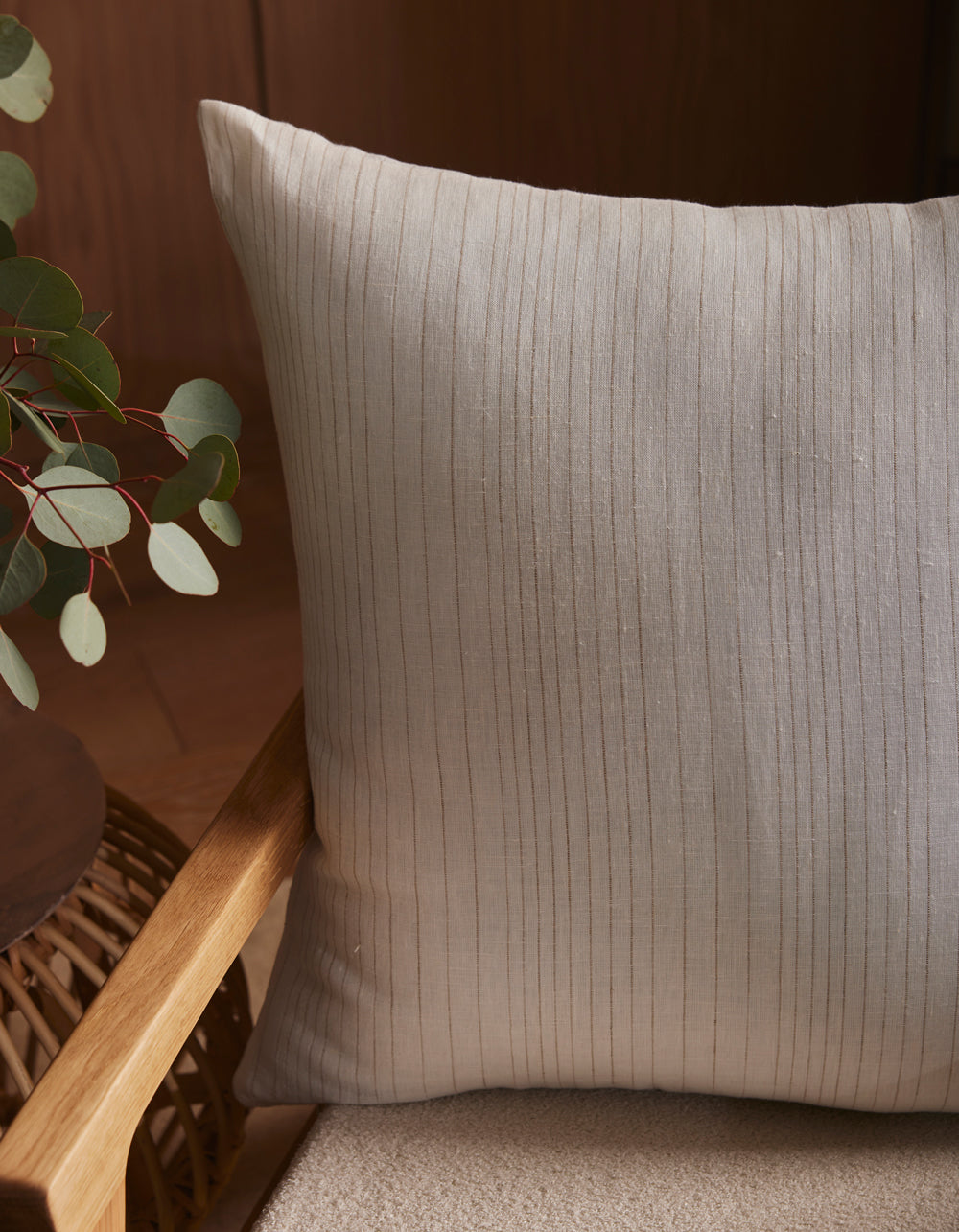 A close-up of the Cozy Earth Striped Throw Pillow, featuring light gray vertical stripes, placed on a beige cushioned chair beside a wooden table with green eucalyptus leaves.