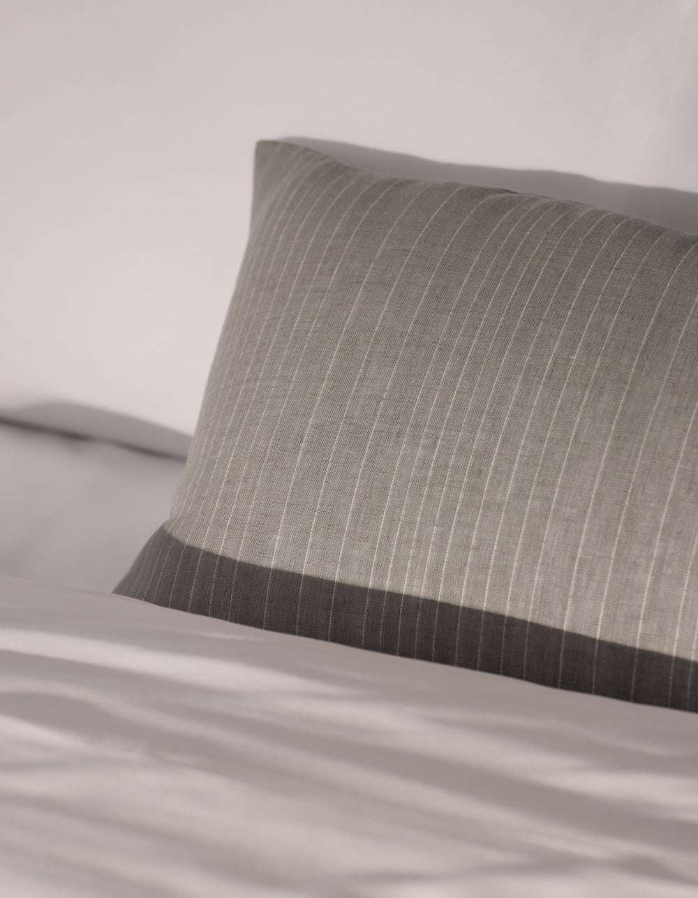 The Cozy Earth Striped Throw Pillow in gray, featuring subtle vertical stripes, rests on a neatly made bed with white sheets, while soft natural light accentuates its texture and premium fabric.