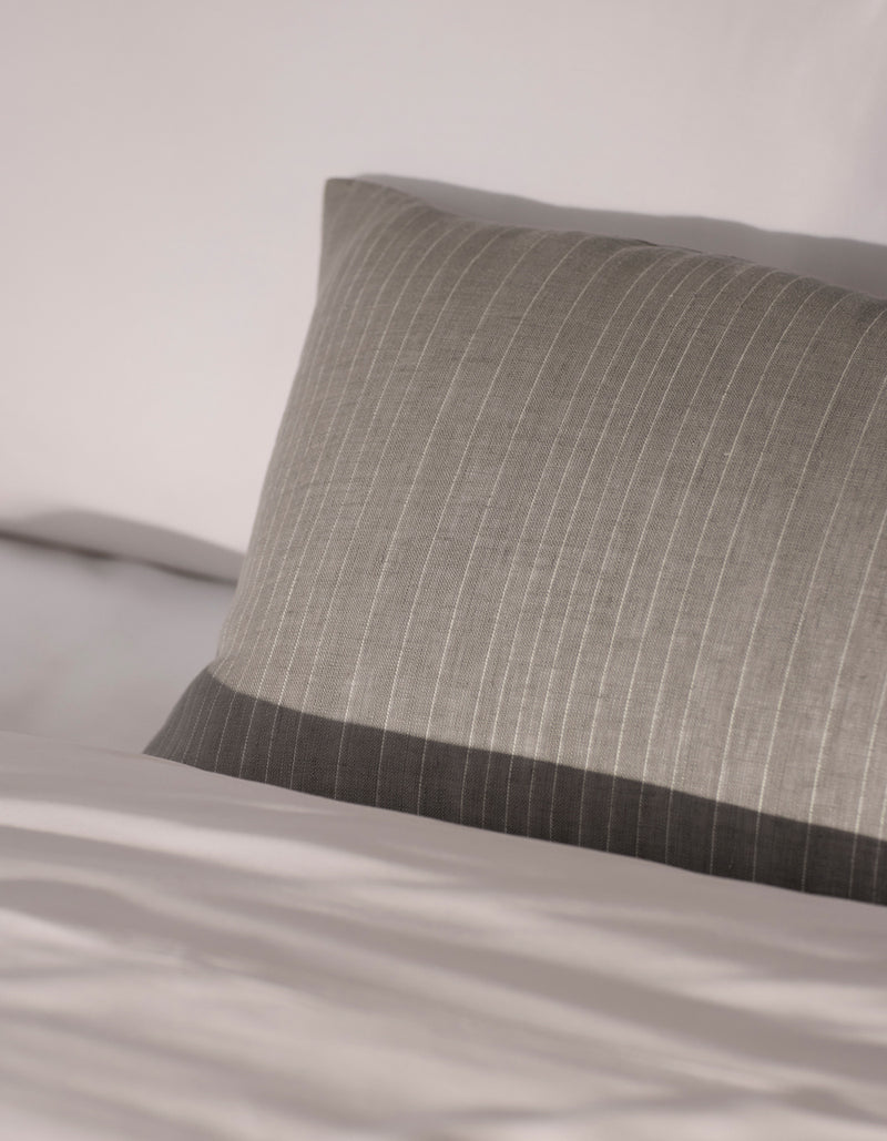 The Cozy Earth Striped Throw Pillow in gray, featuring subtle vertical stripes, rests on a neatly made bed with white sheets, while soft natural light accentuates its texture and premium fabric.