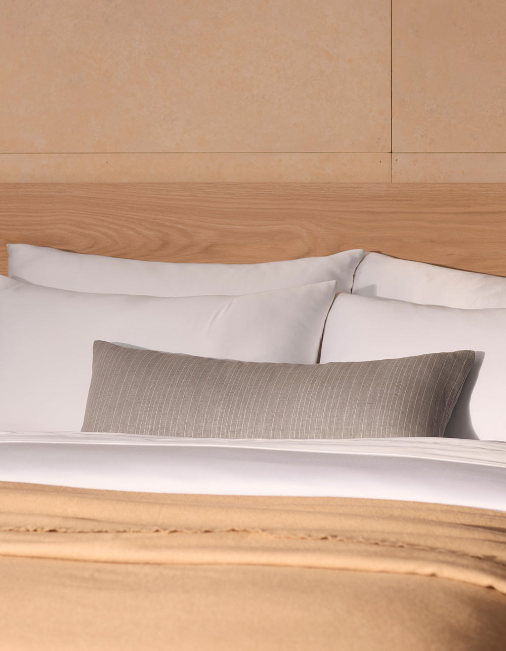 A neatly made bed features Cozy Earth’s Striped Throw Pillow with white pillows, a beige blanket, and a gray cushion, set against a light wood headboard and cream-colored wall.