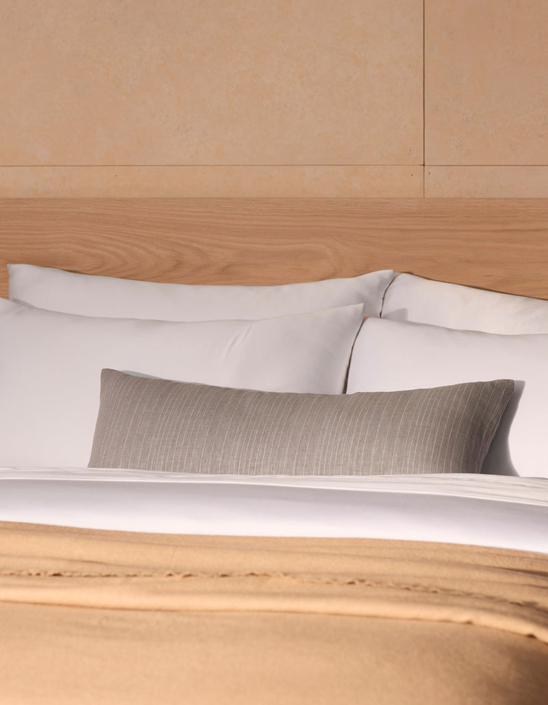 A neatly made bed features Cozy Earth’s Striped Throw Pillow with white pillows, a beige blanket, and a gray cushion, set against a light wood headboard and cream-colored wall.