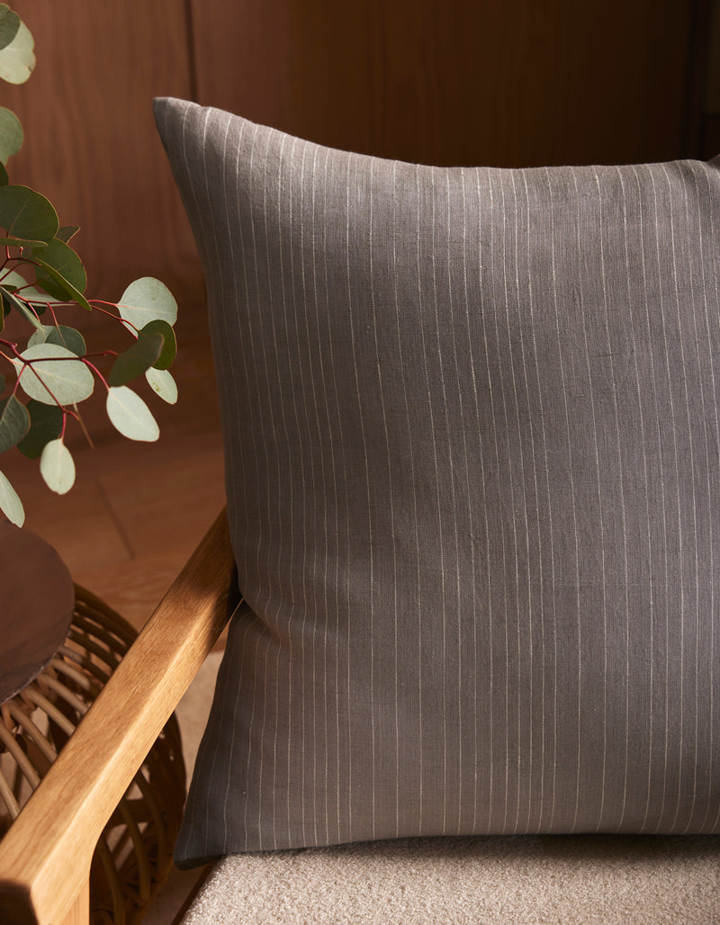 The Striped Throw Pillow by Cozy Earth, featuring thin white pinstripes on gray, sits on a light beige seat. A wooden chair and green plant are nearby, while warm indoor lighting adds to the cozy atmosphere.