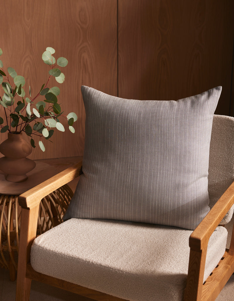 A wooden armchair with a light beige cushion features the Striped Throw Pillow by Cozy Earth and sits by a small table with a brown vase of eucalyptus, set against a wooden wall.