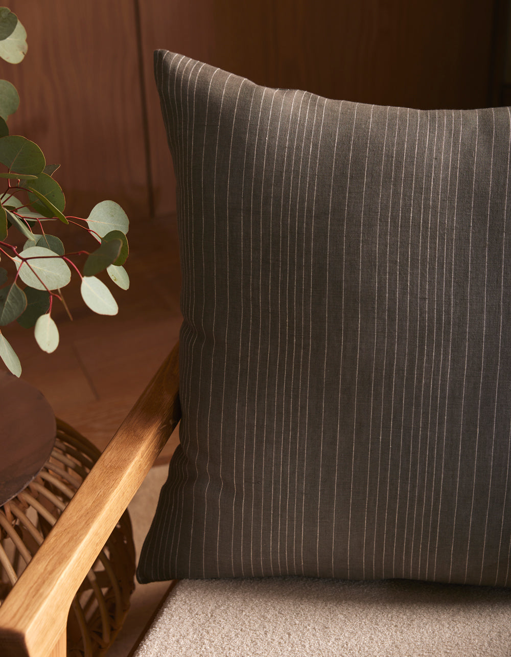 A Cozy Earth Striped Throw Pillow in dark gray with thin white stripes sits on a beige cushioned wooden chair beside a round table decorated with green eucalyptus leaves.