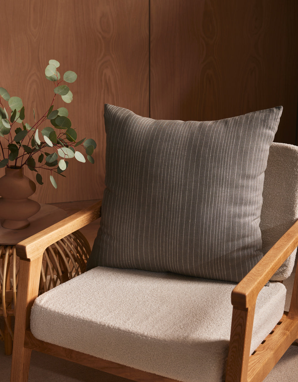 A wooden armchair with a textured, light beige cushion and the Cozy Earth Striped Throw Pillow sits next to a small table with a brown vase of eucalyptus branches, all set against a wooden wall.