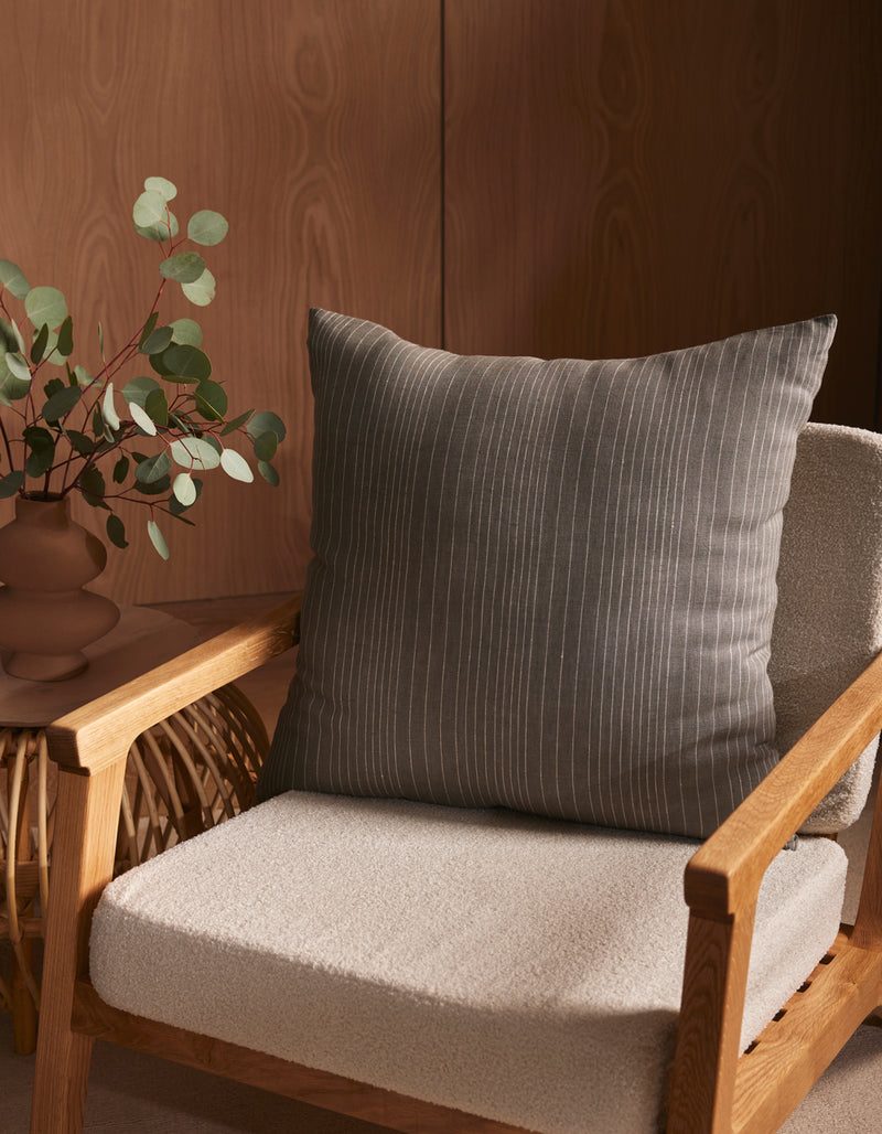 A wooden armchair with a textured, light beige cushion and the Cozy Earth Striped Throw Pillow sits next to a small table with a brown vase of eucalyptus branches, all set against a wooden wall.