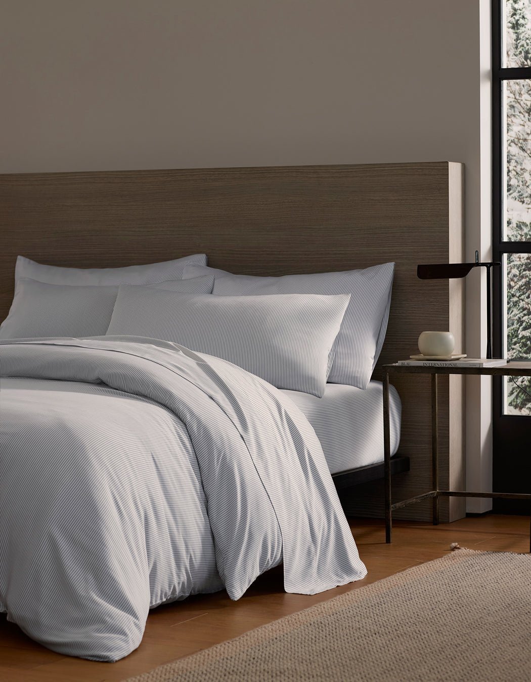 A Bamboo Duvet Cover by Cozy Earth in white covers a cozy bed with pillows against a wooden headboard. A small side table with a dark lamp and books sits nearby, while a large window reveals a snowy outdoor view.