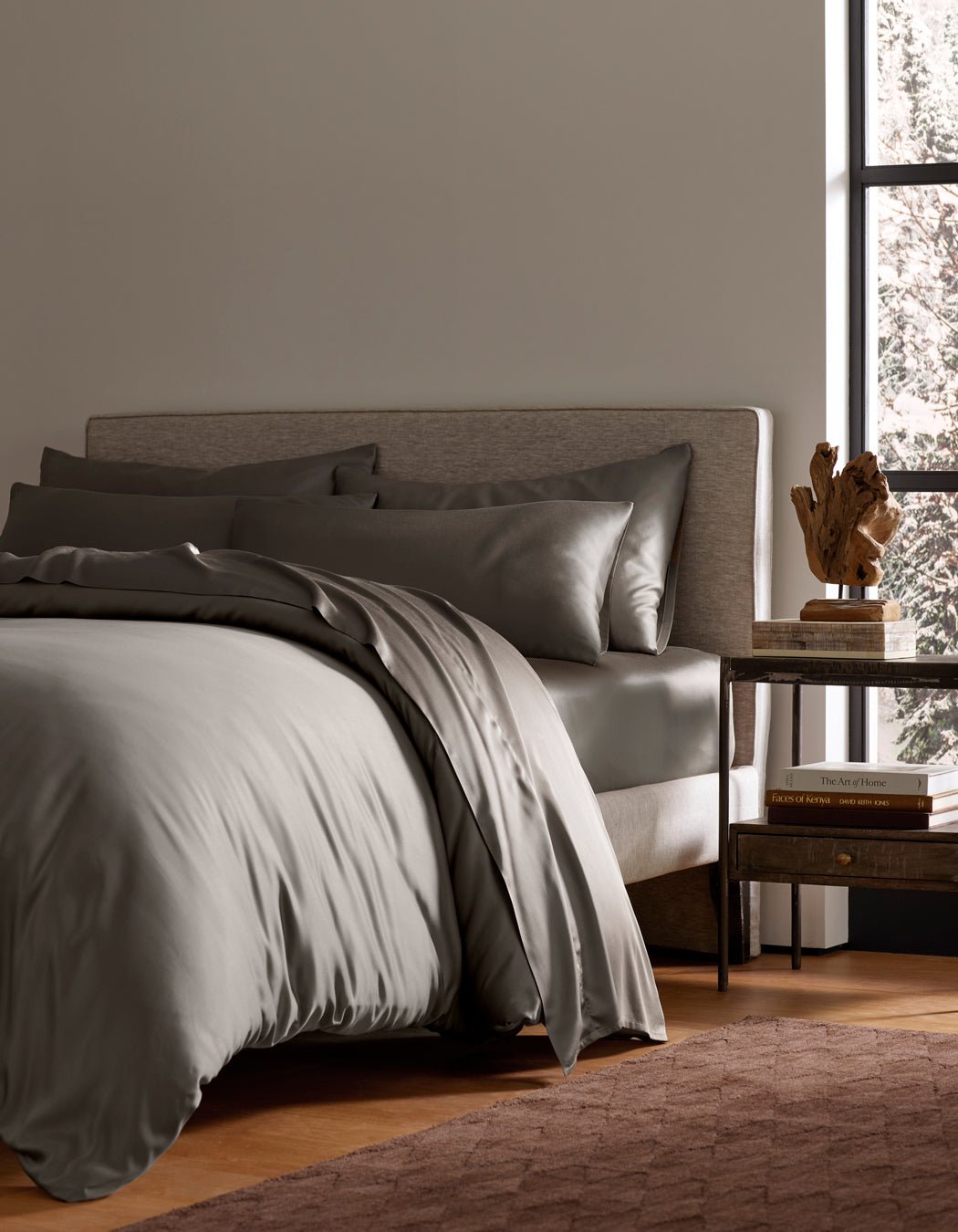A modern bedroom with a neatly made bed featuring Cozy Earth's Bamboo Bedding Ultimate Bundle, two pillows, a beige upholstered headboard, a wooden nightstand with books and a sculpture, and a large window letting in natural light.