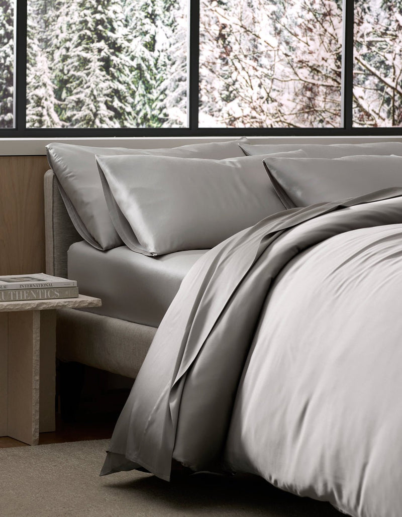 A bed dressed in Cozy Earth’s Bamboo Bedding Ultimate Bundle in light gray sits by a window with a snowy forest view, while a small bedside table holds a stack of books.