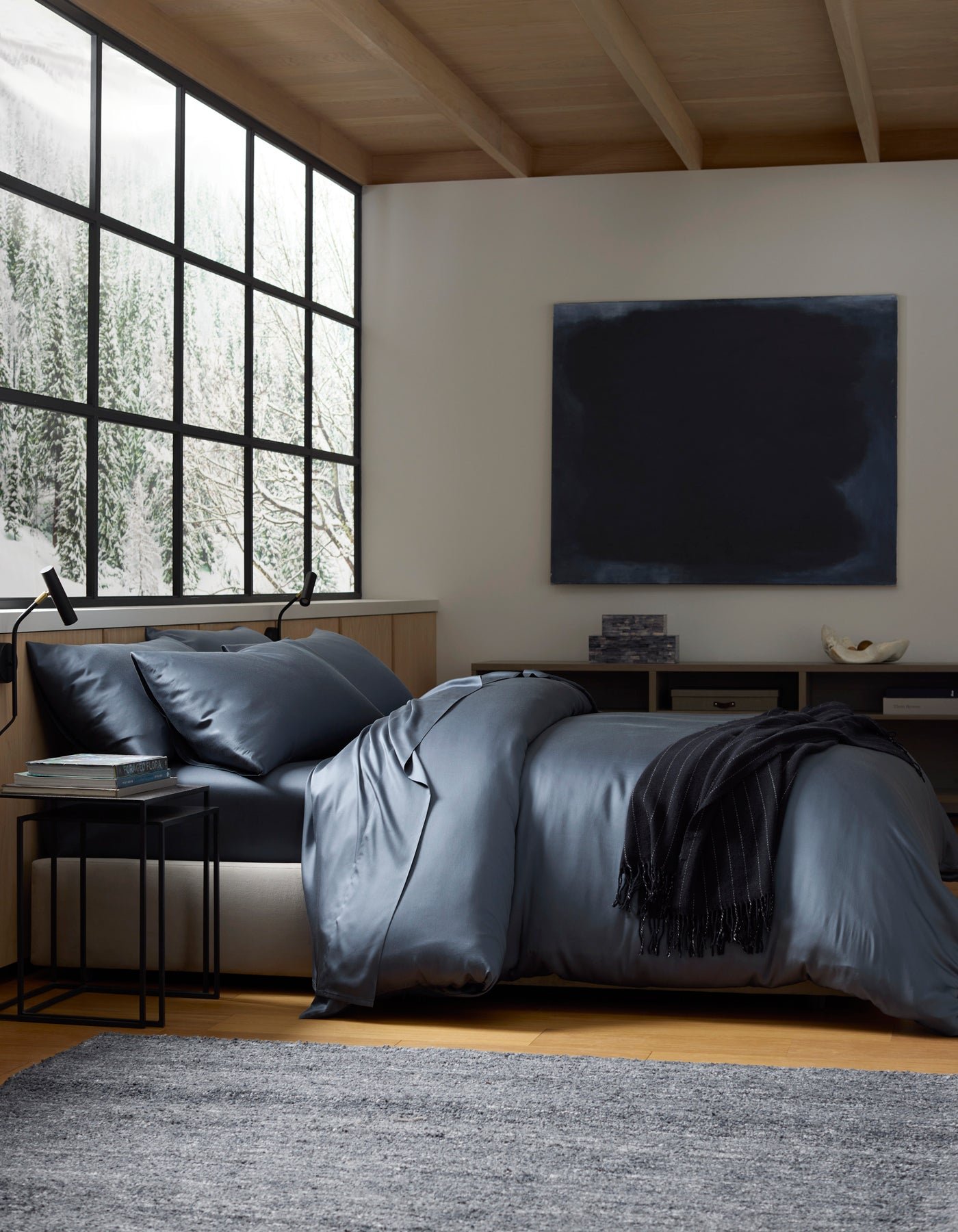 A modern bedroom with a large window showing snowy trees, a bed made with Cozy Earth's Bamboo Bedding Deluxe Bundle in dark grey, a black throw, side table, grey rug, and a dark abstract painting on the wall.