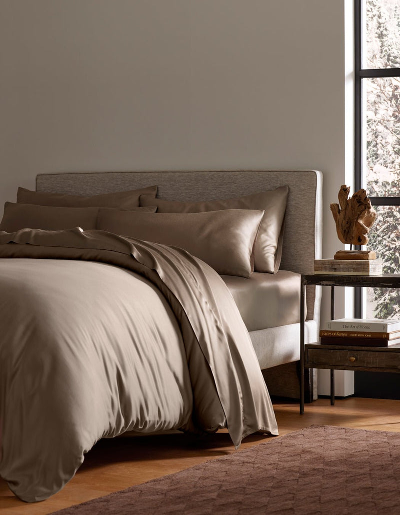 A modern bedroom featuring the Cozy Earth Bamboo Bedding Ultimate Bundle on a neatly made bed, with a gray upholstered headboard, dark wooden nightstand with books and sculpture, and a large window letting in natural light.