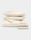The Bamboo Bedding Ultimate Bundle by Cozy Earth, shown on a light grey background, features a neatly folded cream-colored duvet, two pillows, and a stack of sheets.|Color:Creme
