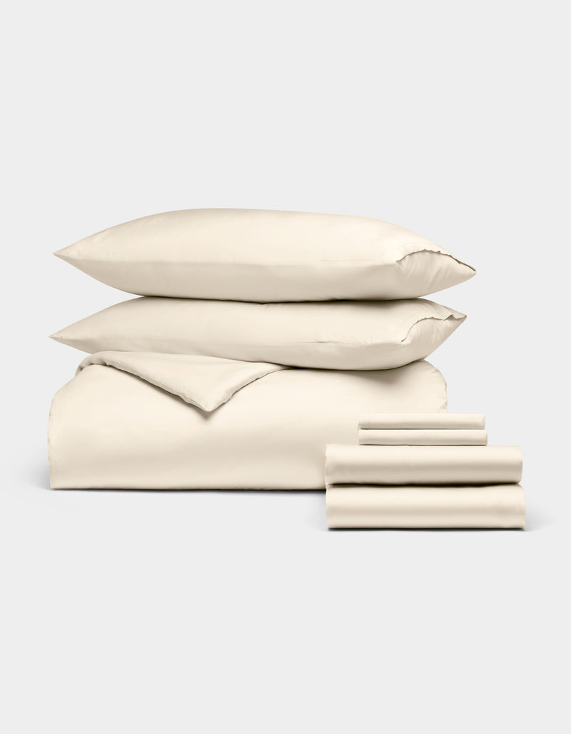 The Bamboo Bedding Ultimate Bundle by Cozy Earth, shown on a light grey background, features a neatly folded cream-colored duvet, two pillows, and a stack of sheets.