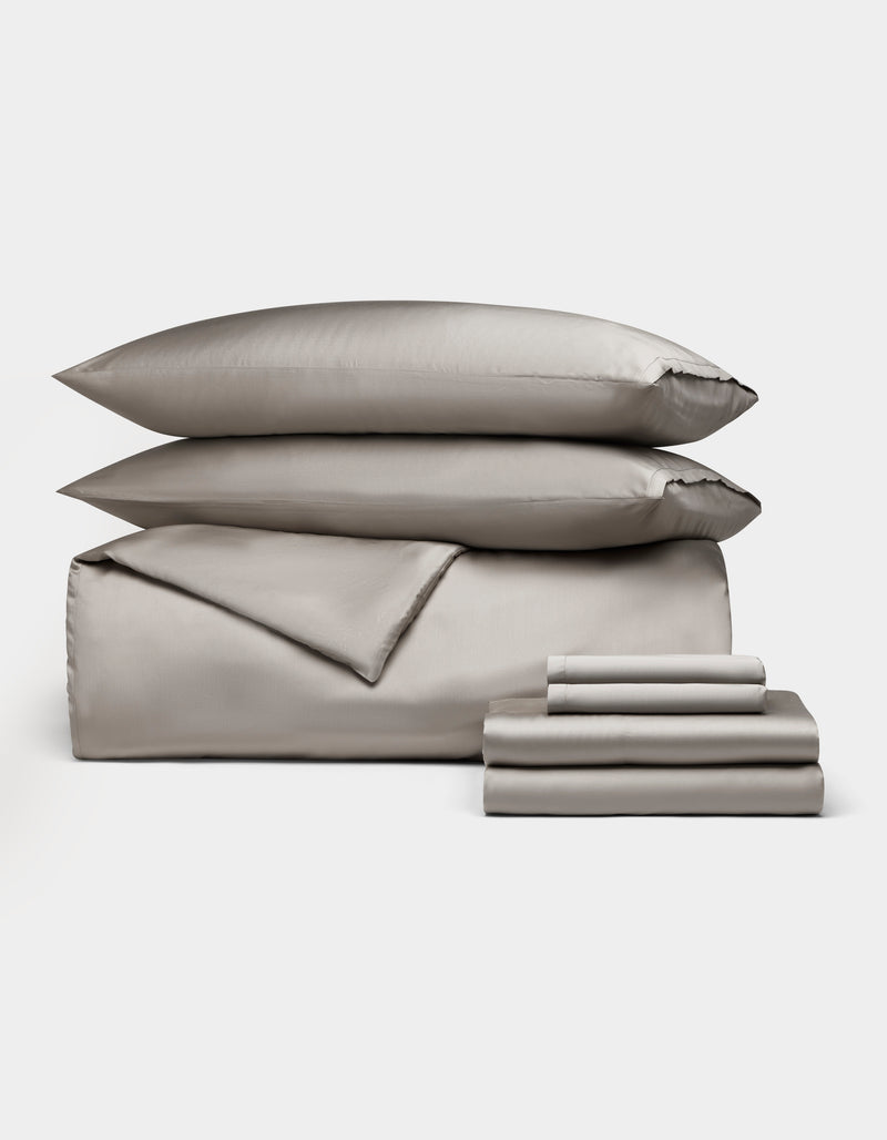 The Bamboo Bedding Ultimate Bundle by Cozy Earth features neatly stacked gray bed linens, including two pillows, a duvet, and folded sheets, all displayed against a plain white background.
