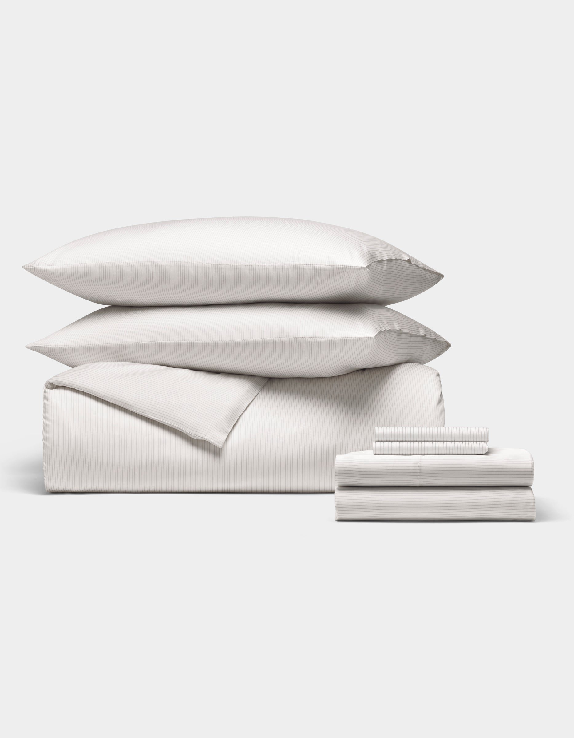 The Cozy Earth Bamboo Bedding Ultimate Bundle features two pillows, a folded comforter, and a stack of sheets and pillowcases in white stripes, all arranged neatly against a plain light background.