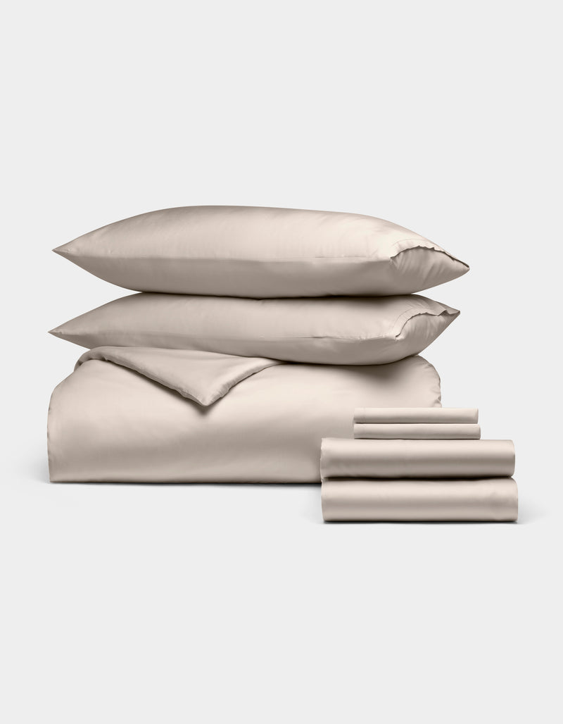 The Cozy Earth Bamboo Bedding Ultimate Bundle, shown in light beige on a plain white background, features two stacked pillows, a folded duvet, a flat sheet, and two folded pillowcases arranged neatly.