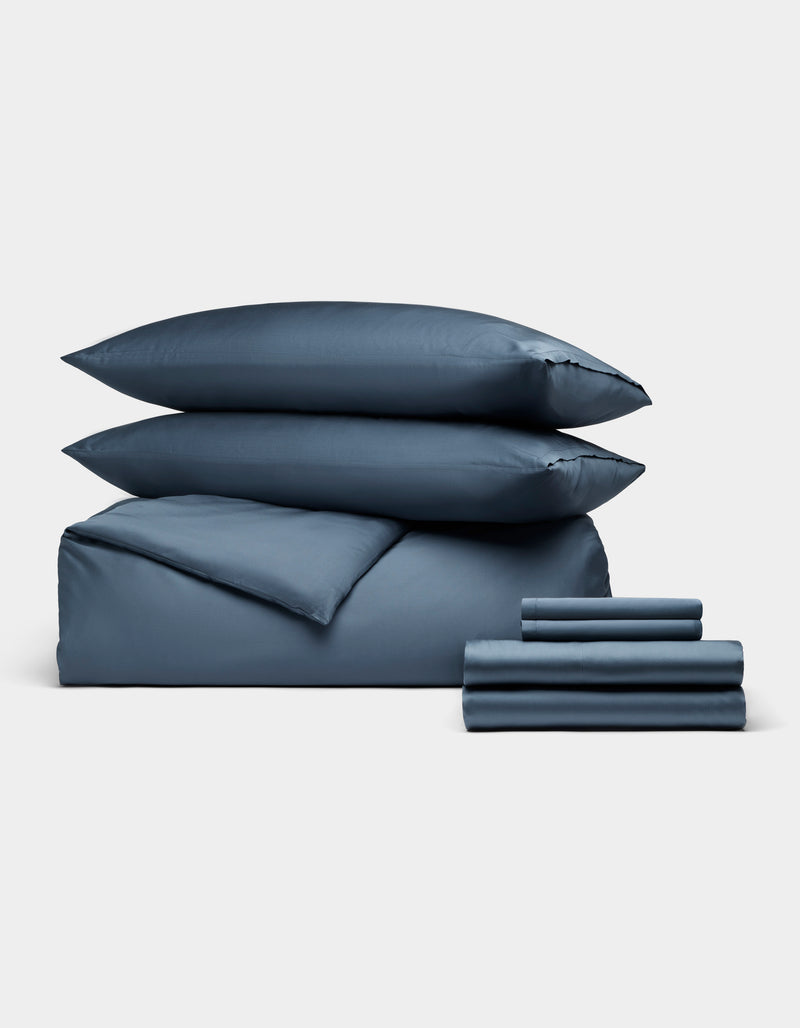 The Cozy Earth Bamboo Bedding Ultimate Bundle features a neatly stacked set of dark blue bedding—two pillows, a folded duvet, and several sheets—all displayed against a plain light gray background.